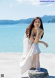 A woman in a white dress sitting on a rock by the water.